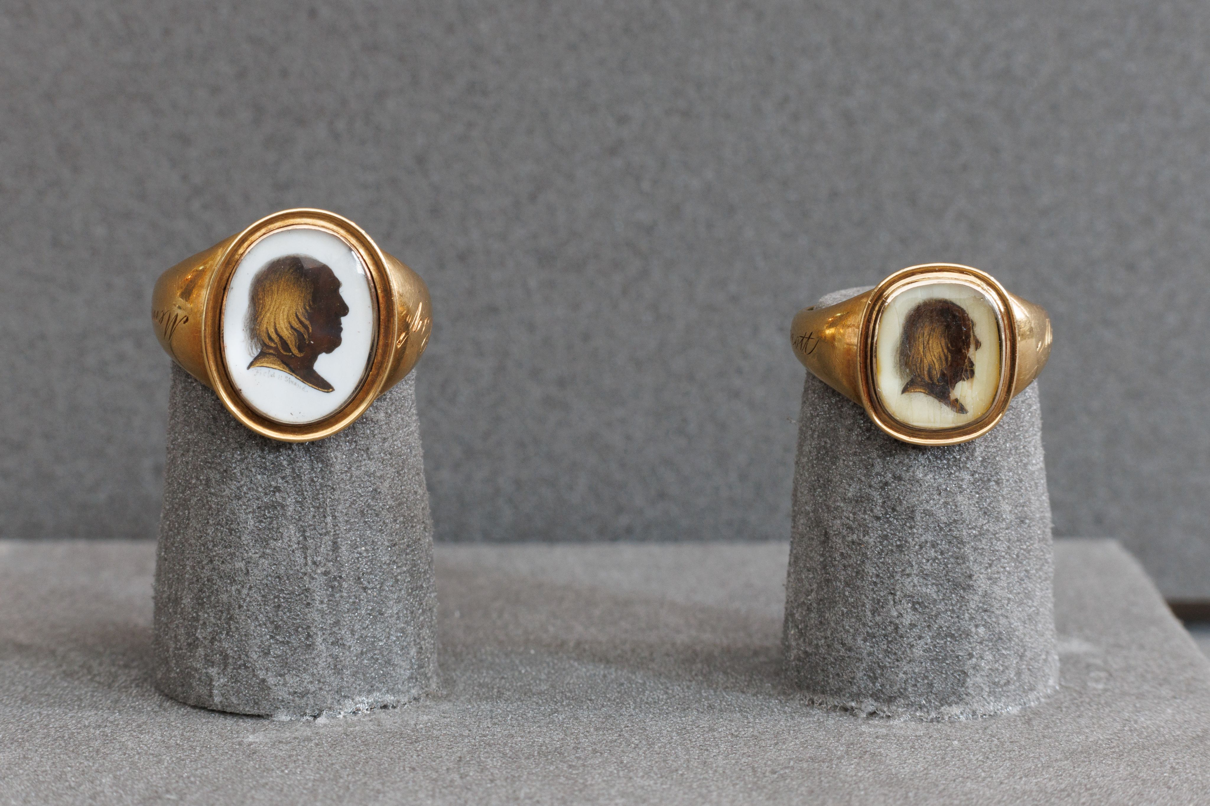 Image shows two gold rings on a grey surface. The left ring has a silhouette profile, and the right ring has an aged portrait. 